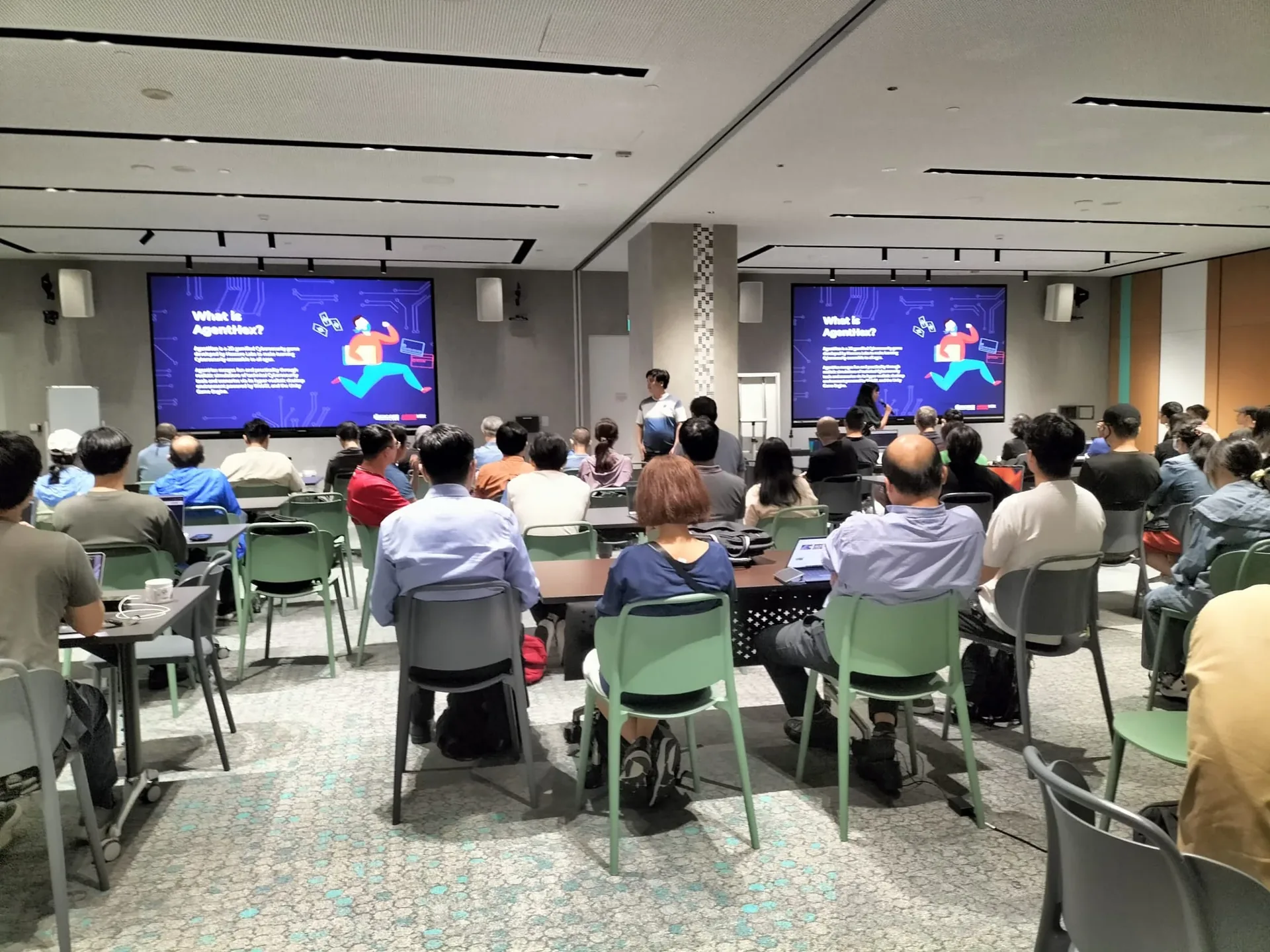 Adult learners seated in rows facing two large screens displaying cybersecurity content at an indoor workshop event