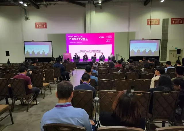 Audience members seated in an auditorium facing a stage with a bright pink screen displaying a tech talk session at the IMDA Tech Skill Up Festival