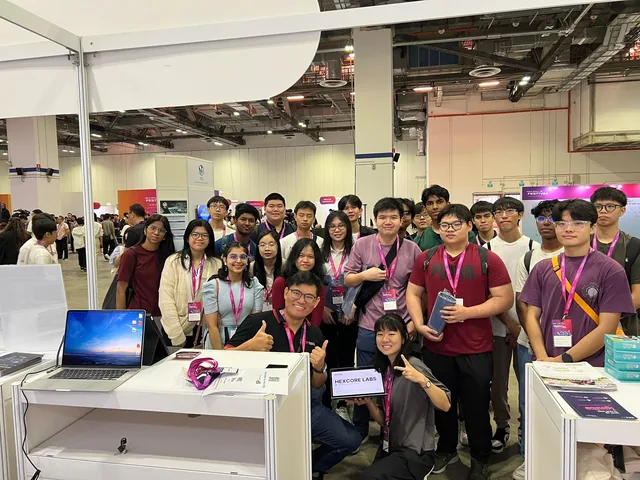 Group of students and professionals wearing lanyards posing together at an indoor event booth with a laptop and materials on display
