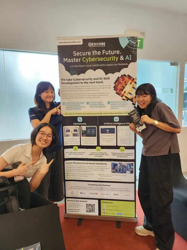 Three women posing beside a Hexcore Labs exhibition poster about cybersecurity and AI training at the IDE Series 2025 competition
