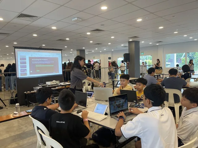 Students seated at tables with laptops during a workshop at the IDE Series 2025 competition, with a presenter at a screen in the background