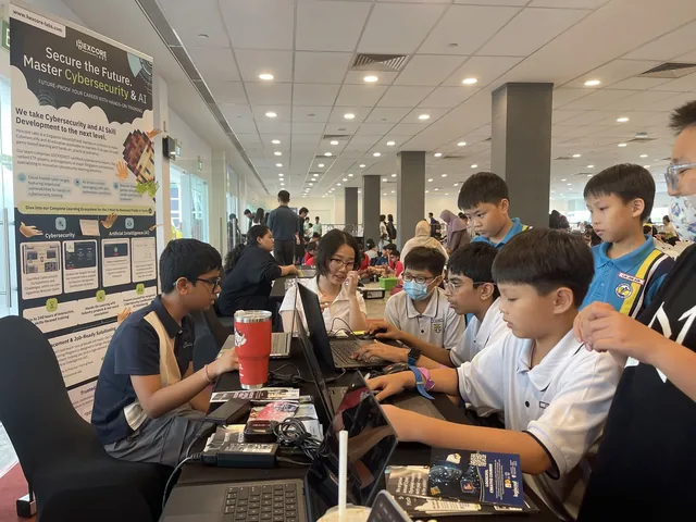 Students gather at an exhibition booth where presenters demonstrate technology projects, with informational posters displayed on the wall behind them