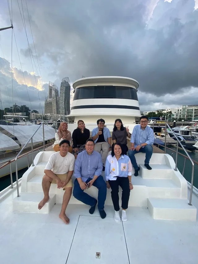 Eight people posing together on the deck of a white yacht moored in a marina with city buildings and cloudy sky in the background