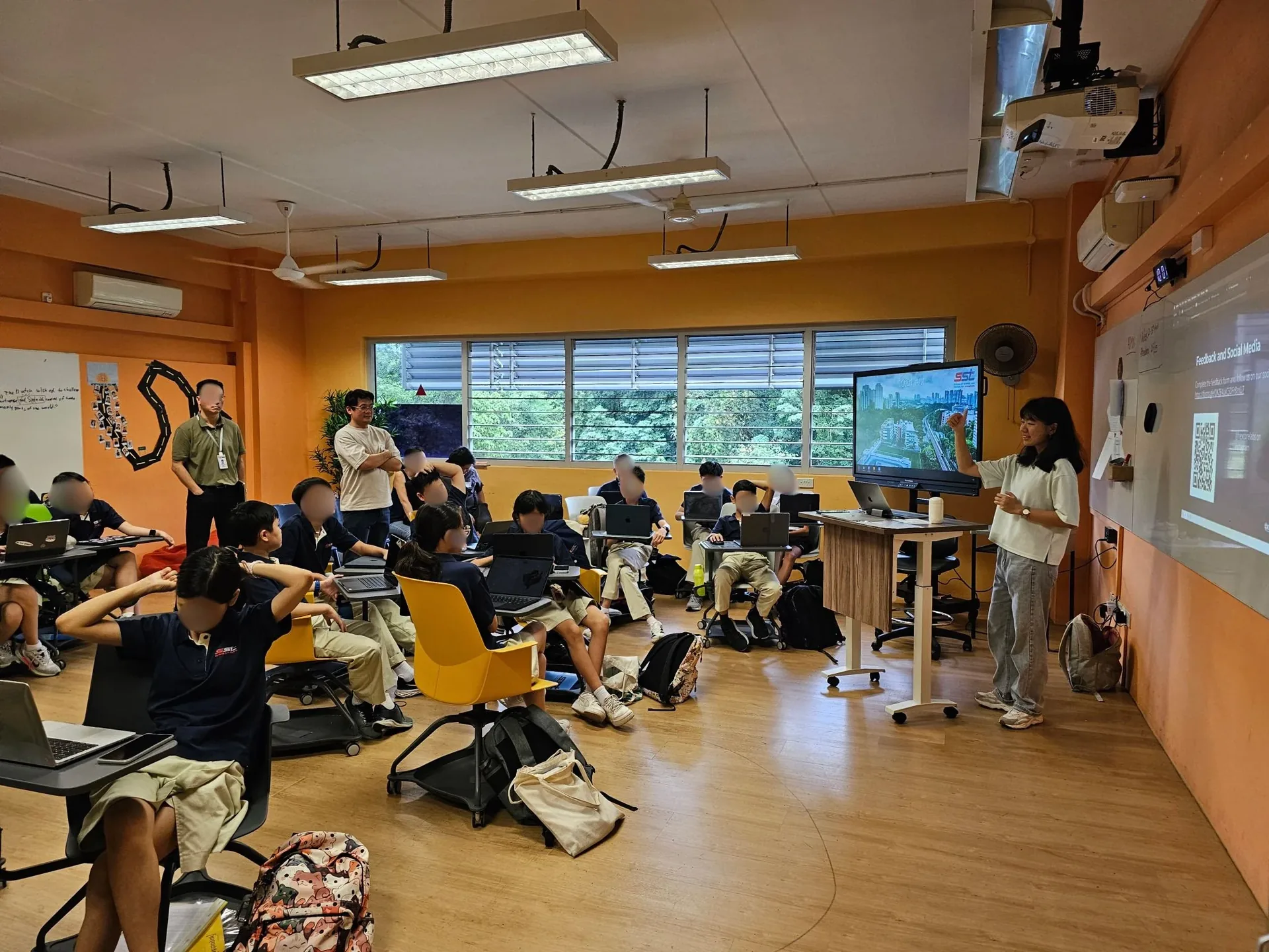 Students seated in a bright orange classroom with large windows, engaged in a cybersecurity learning session with instructors present