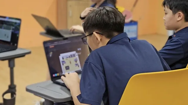 Students in school uniforms work on laptops during a cybersecurity lesson in a classroom with yellow walls
