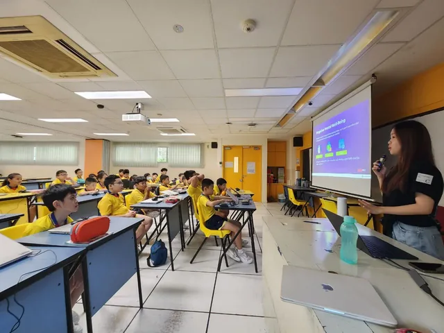 A woman presents to a classroom of students wearing yellow shirts seated at blue desks, with a projection screen visible on the right wall