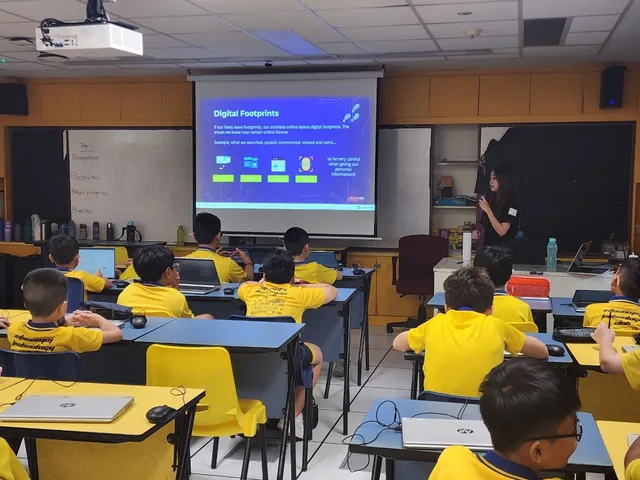 Students in yellow shirts sit at desks in a classroom while an instructor presents a slide about Digital Footprints on a projected screen