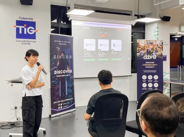 A presenter holds a microphone while speaking to an audience at an indoor cybersecurity meetup, with a projected screen display and Division 0 banners visible