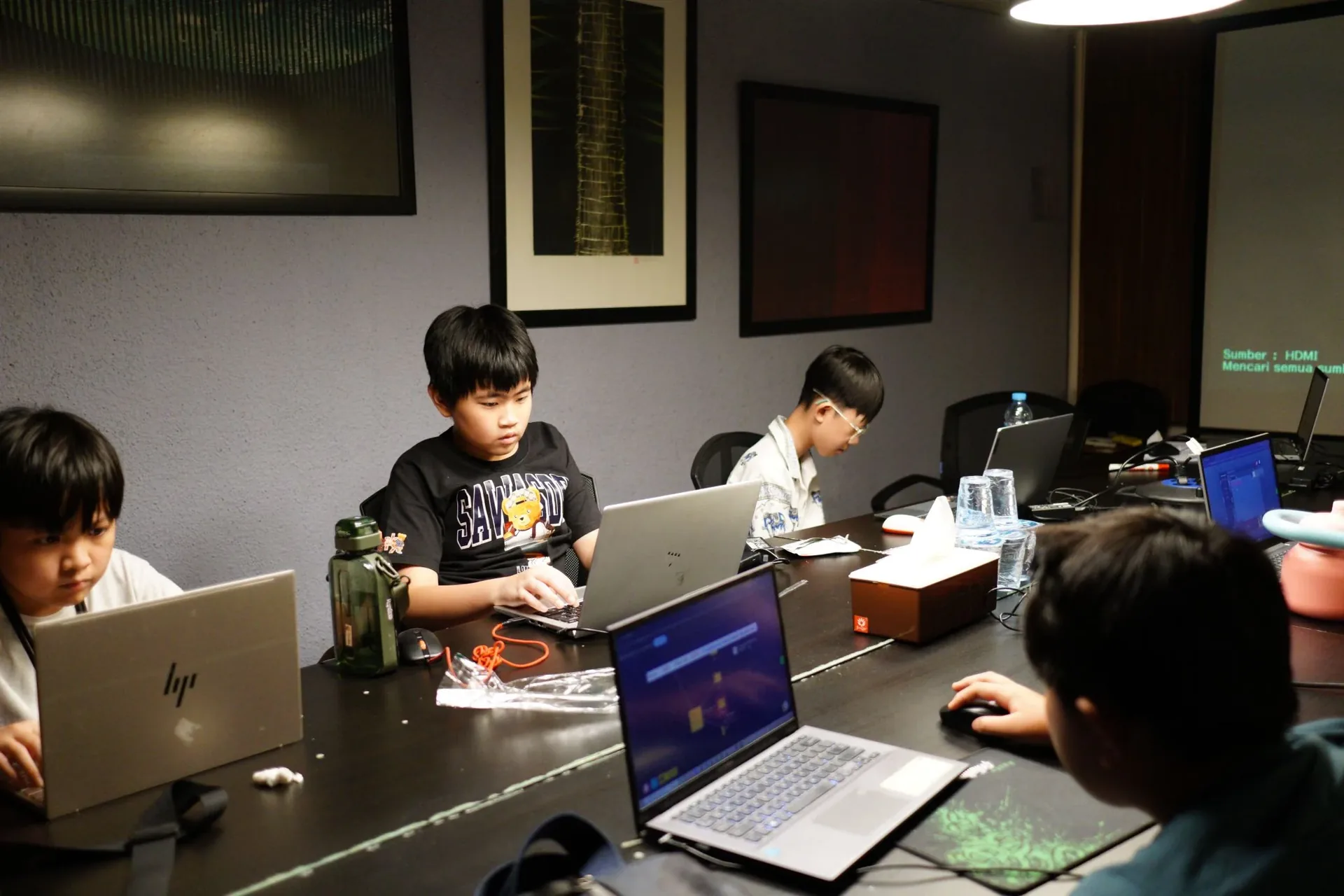 Children aged 8–16 working on laptops at a table during a cybersecurity holiday camp workshop