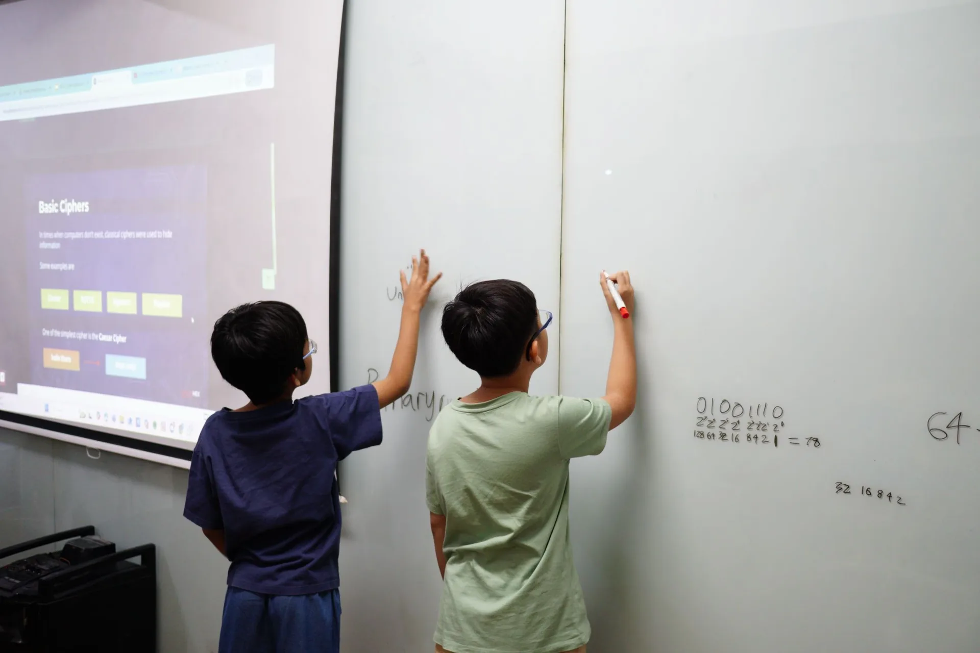Two children aged 8–16 stand at a whiteboard with a projected screen displaying cybersecurity content during the Cyber Kids Adventure Holiday camp