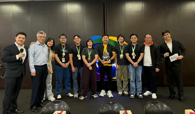 Group of approximately 12 people standing in a line indoors, including students wearing lanyards and medals, alongside adults in business attire, at the Hexcore