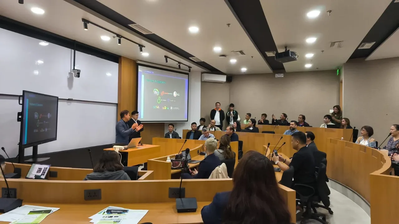 Students and judges seated in a tiered lecture hall during a competition event, with a presenter at a podium and projection screen displaying content