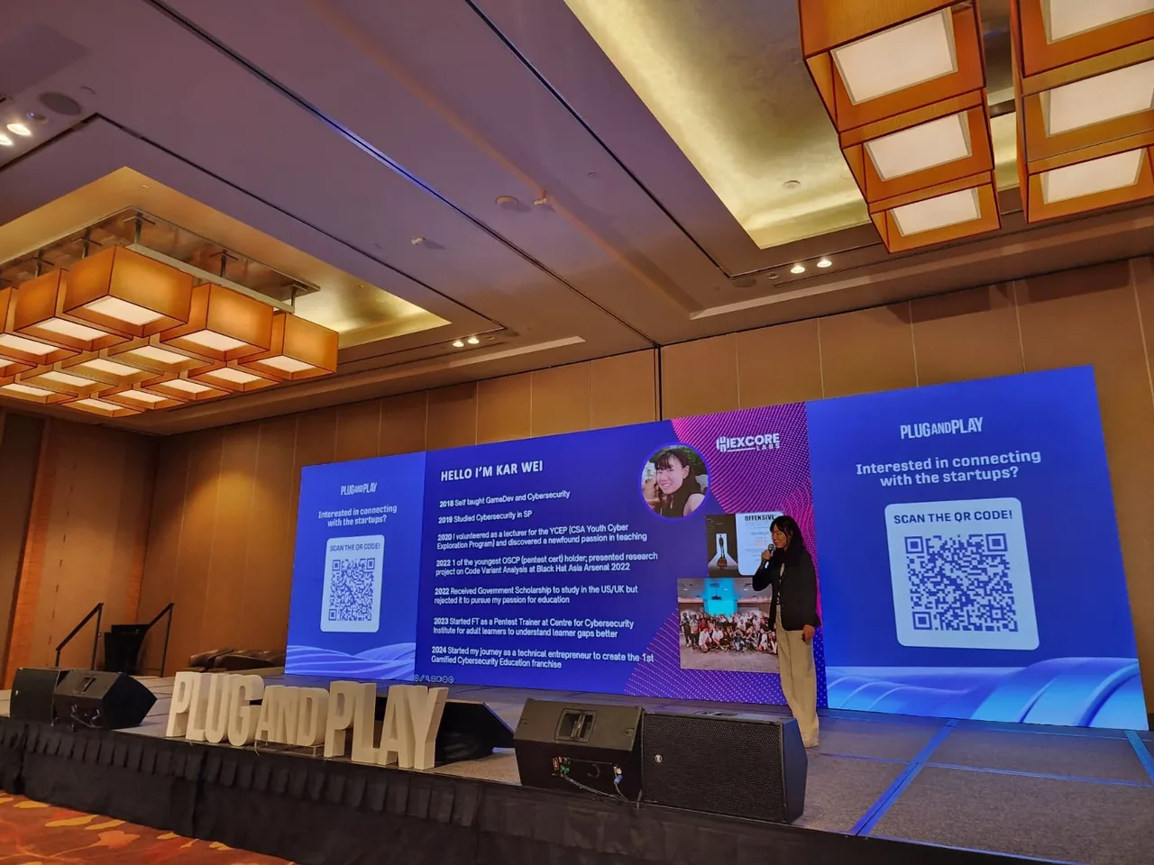 A person stands at a podium on stage at the Plug and Play APAC Summit 2024, delivering a pitch in front of large blue display screens