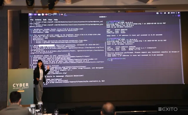 Presenter standing on stage beside a large screen displaying code during a live hacking demonstration at a cybersecurity event