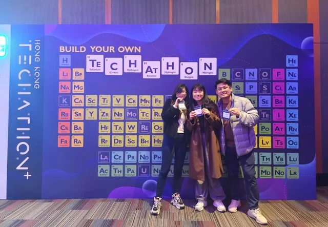 Three people posing in front of a purple backdrop displaying a periodic table and "BUILD YOUR OWN" and "HKSTP TECATHON+" text at Hong Kong Science and Tech Park