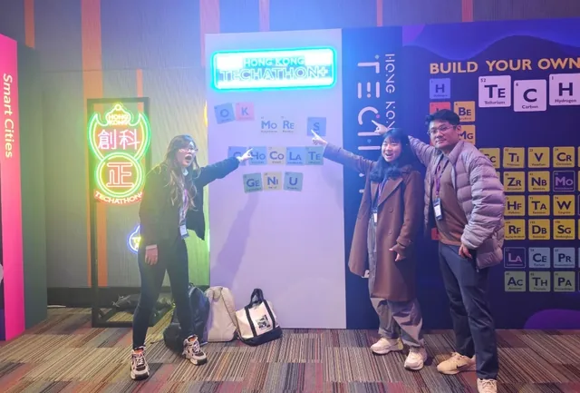 Four people posing at the Hong Kong Science and Tech Park event, standing in front of colourful neon signs and periodic table displays
