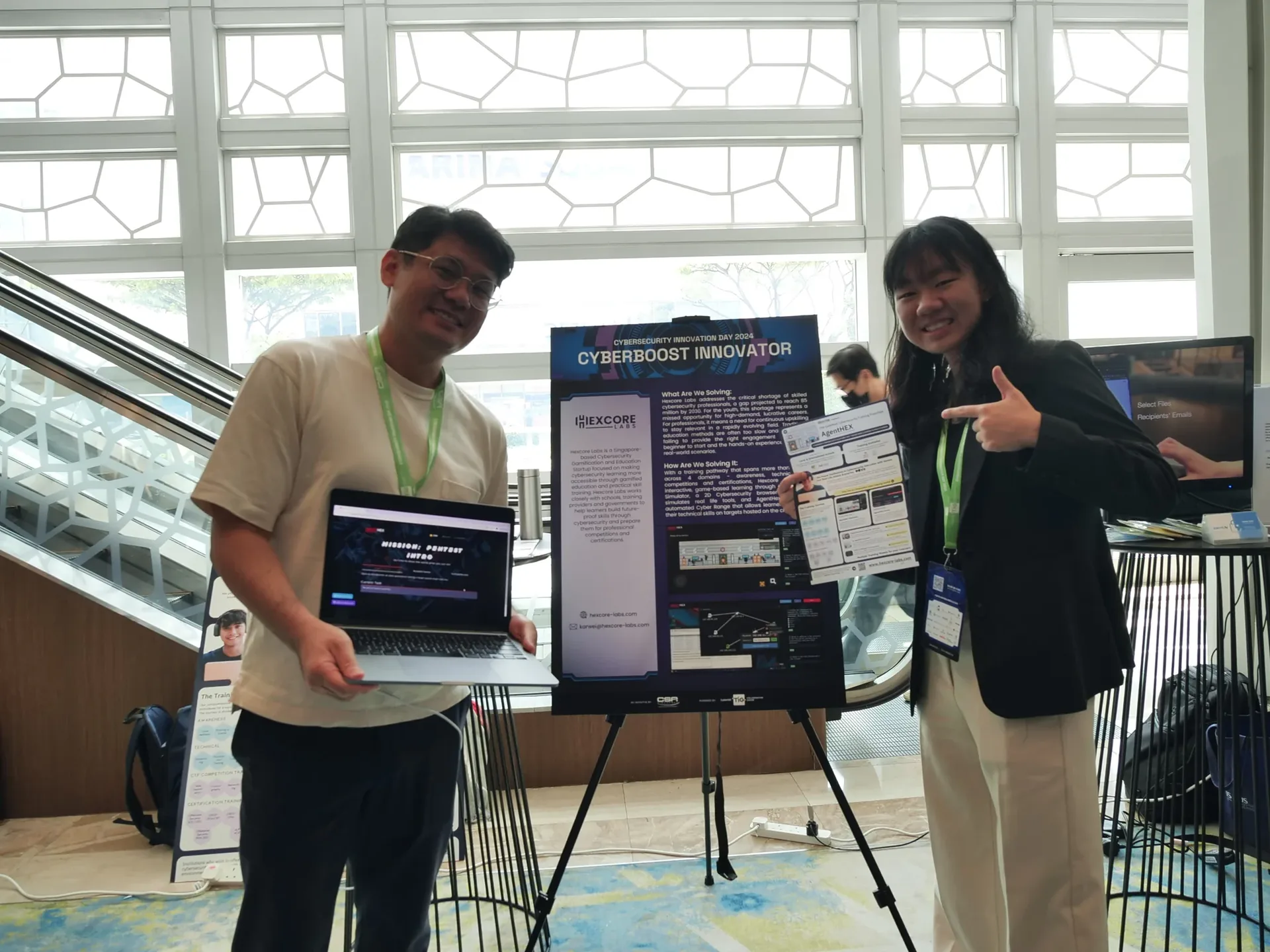 Two people with green lanyards stand beside a display board titled "Cyberboost Innovator" at an indoor exhibition venue with large windows