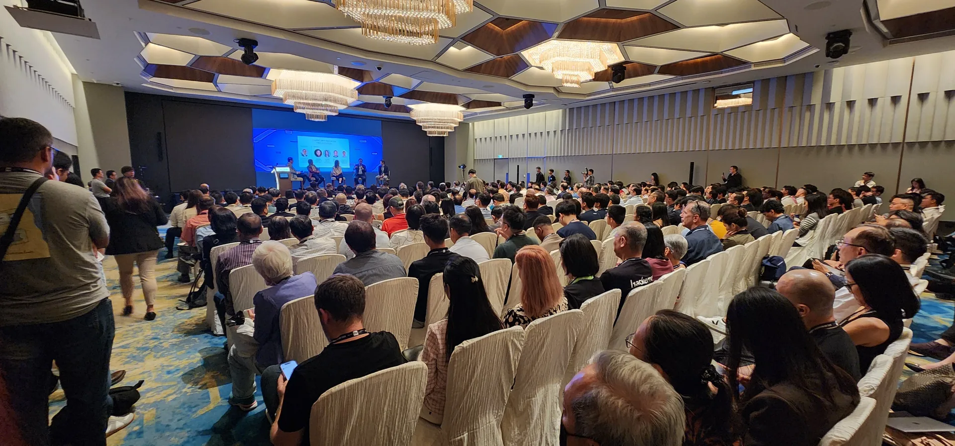 Large audience seated in a conference hall facing a stage with blue lighting during the Cybersecurity Innovation Day 2024 Final Demo Day