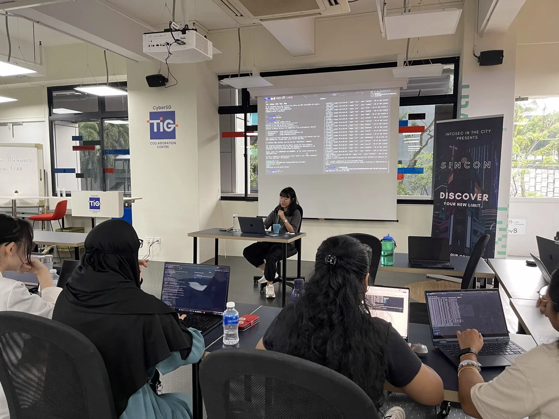 Women participants seated at laptops in a workshop room, facing a presenter at a desk with code displayed on a large screen behind her