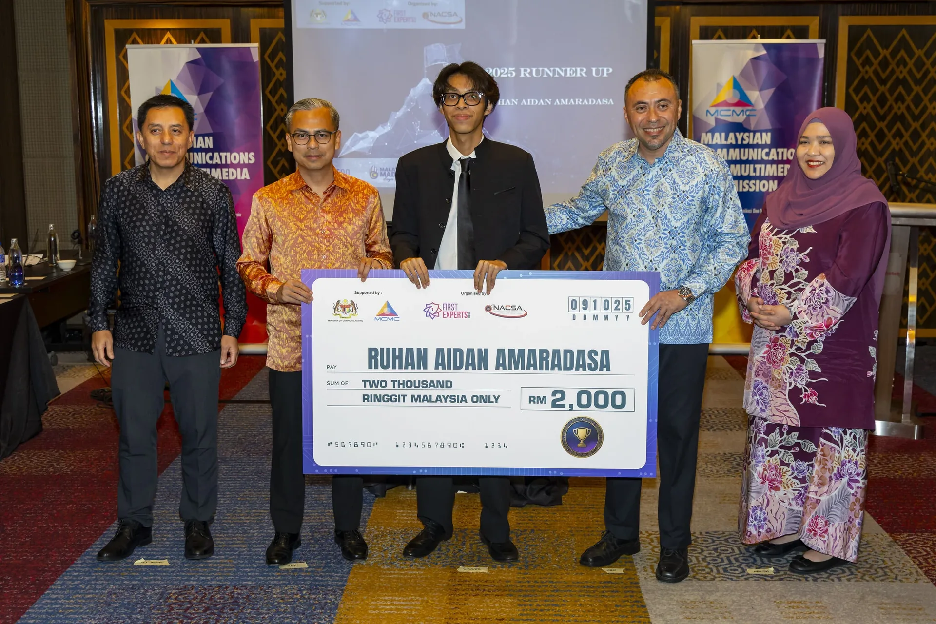 Five people holding a large cheque for two thousand ringgit at an indoor event with logos and banners visible in the background