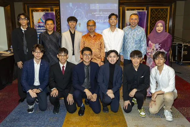 Group photo of twelve people in formal and traditional attire at an indoor event, arranged in two rows with a Malaysia Day banner visible behind them