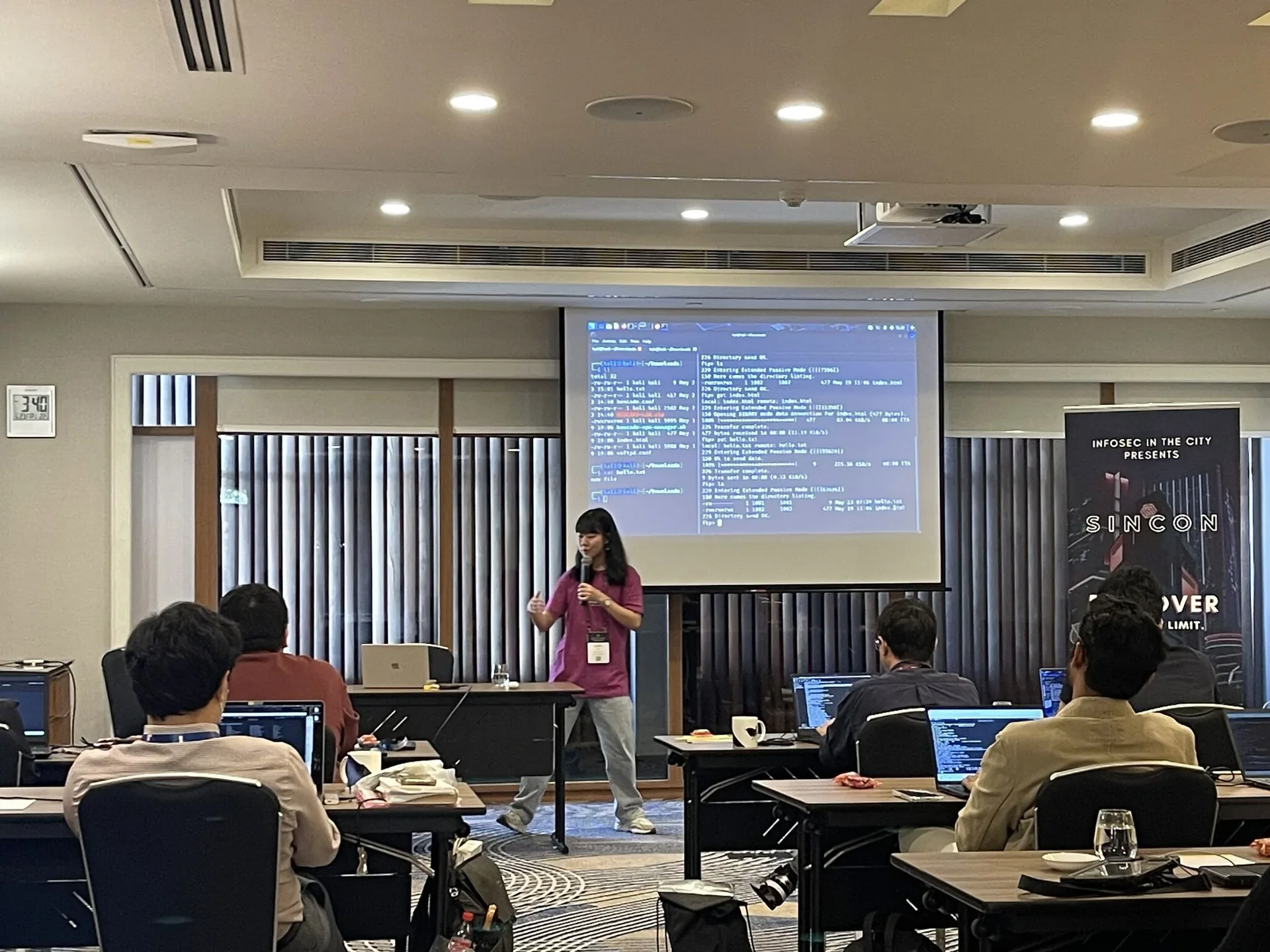 Presenter leading a hands-on cybersecurity workshop in a conference room, with code displayed on a large screen behind them and participants seated at tables wi